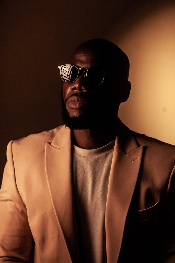 Portrait of Surinamese artist P_ton (Garry Waterberg) wearing sunglasses and a beige blazer in warm, dramatic light